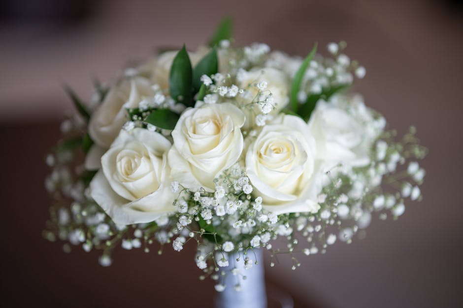 A close-up of a bouquet composed of white roses with tightly curled petals and lush green leaves, accented by delicate sprigs of baby's breath with tiny white flowers. The bouquet is arranged in a com