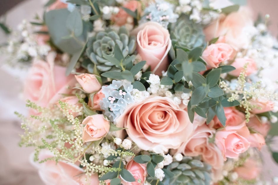 A close-up view of a floral bouquet arranged with large, soft pink roses, complemented by smaller pink spray roses and light blue succulents. The bouquet includes delicate white baby's breath, airy gr