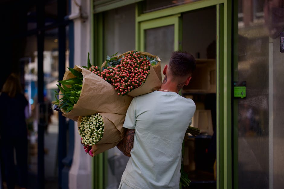 A person with short dark hair, wearing a light-colored T-shirt, is seen from behind holding a large bouquet of fresh flowers wrapped in light brown paper. The bouquet includes pink and white berries,