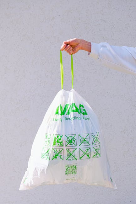A person's hand, dressed in a white sleeve, holds a green handle attached to a white biodegradable recycling bag with green printed recycling symbols and text. The background is a plain, light-colored