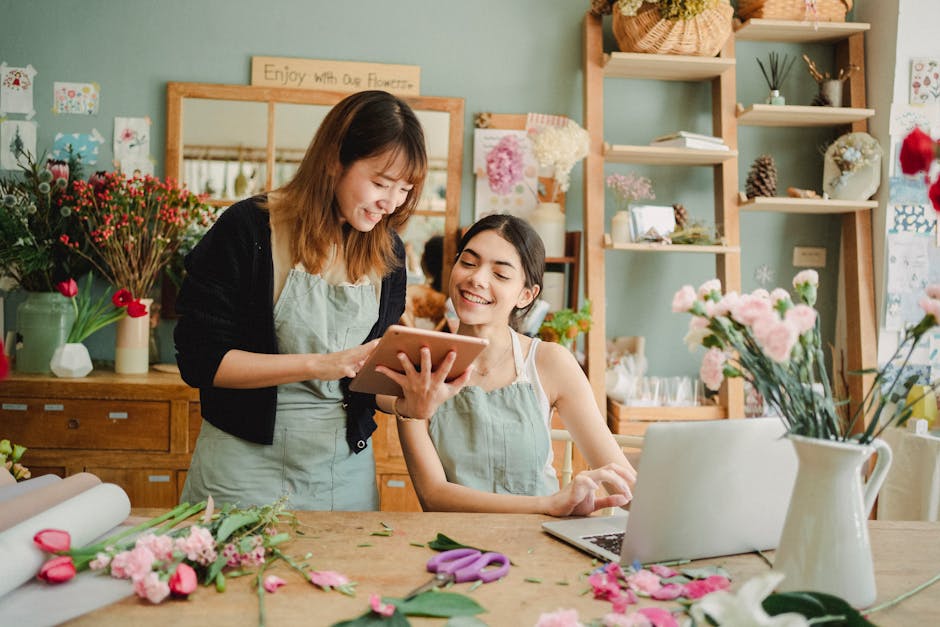 Two women inside a flower shop are engaged in a floral arrangement activity. The woman on the left, with shoulder-length brown hair, is standing and holding a digital tablet, smiling as she shows some