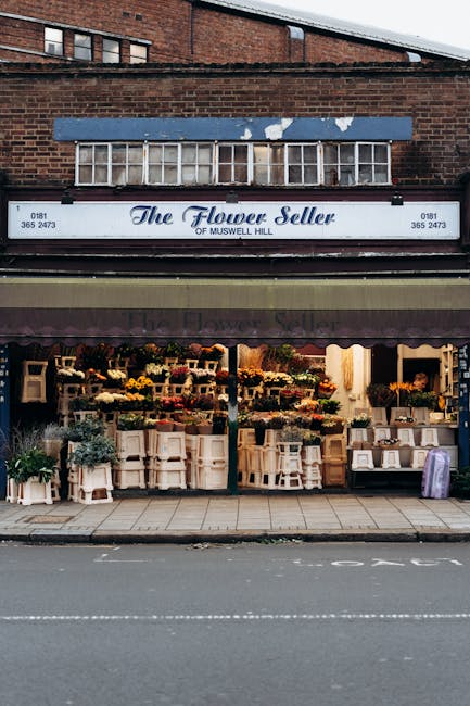 The storefront of The Flower Seller of Muswell Hill, featuring a wide display of floral arrangements and bouquets arranged on tiered white shelves and tables under a dark purple awning with the shop's