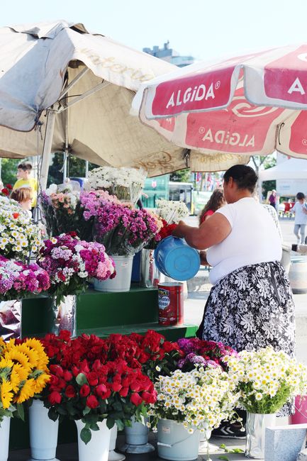 A flower stall at an outdoor market under a large, beige canopy is filled with a variety of fresh floral arrangements. Prominent in the image are vibrant bouquets of chrysanthemums in shades of purple