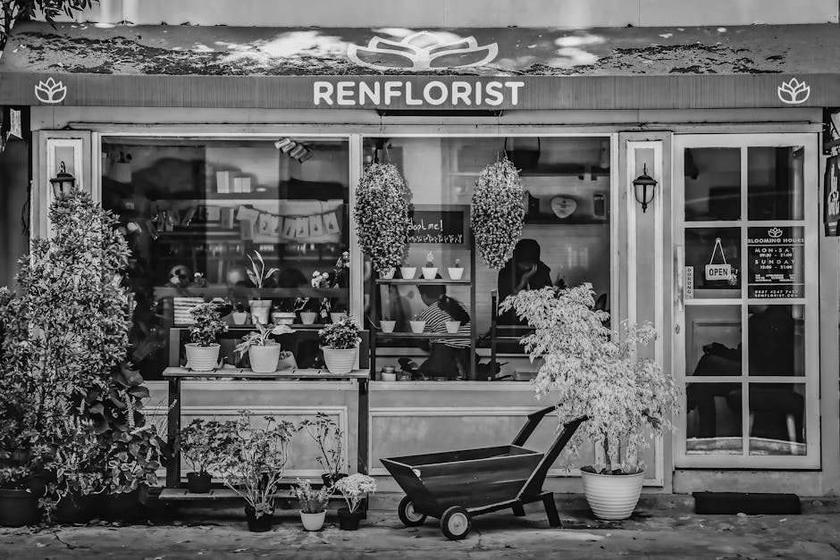 Black and white photograph of the storefront of Ren Florist, a local flower shop with large glass windows displaying an interior filled with floral arrangements and customers. The shop's signage reads