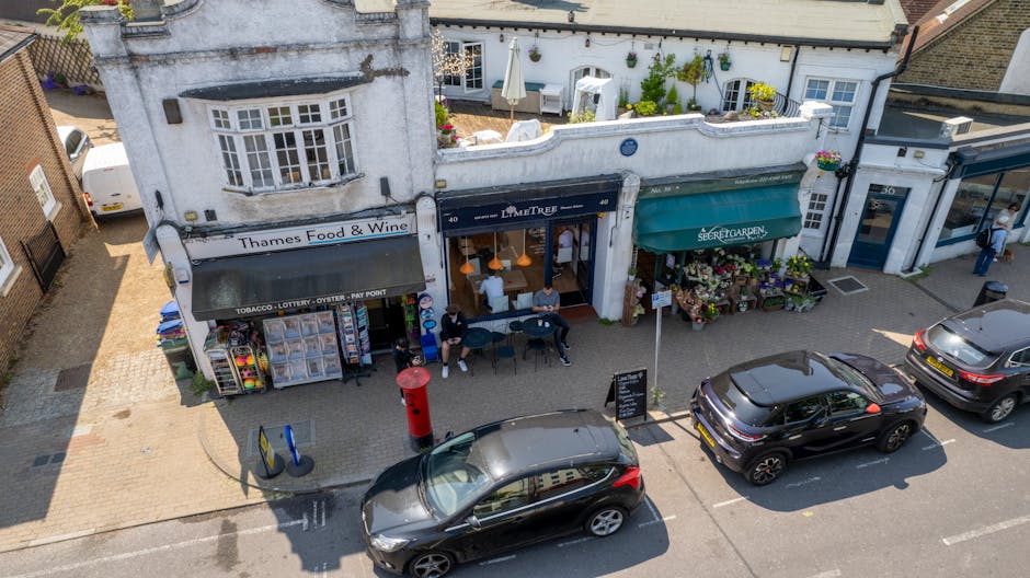 An aerial view of a small row of shops on a street, including a floral shop with a green awning labeled 'Secret Garden', displaying vibrant potted plants and flowers outside. Next to it, a cafe with a