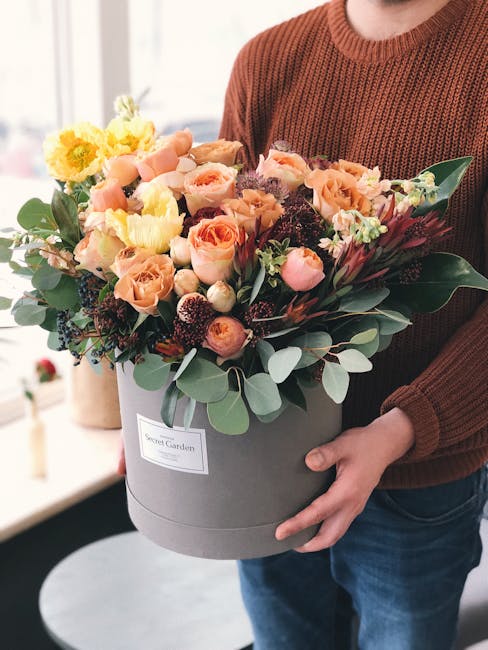 A person wearing a rust-colored knitted sweater is holding a large, round, light grey hatbox-style floral arrangement from Haringey Flowers, featuring a diverse selection of fresh flowers including so
