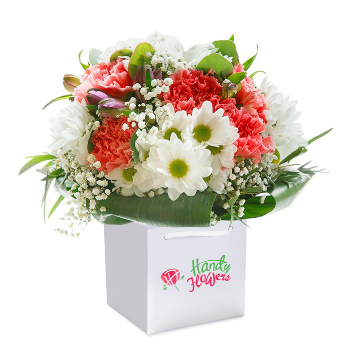 A lush, expertly arranged bouquet sits upright in a spotless square gift bag, echoing the lively charm of Haringey's diverse streets and green spaces. At its core, a delightful mix of soft coral, peach, and vivid red carnations radiates energy, their signature frilled petals delivering rich texture and warmth-a tribute to the colourful scenes at Wood Green and Bounds Green. Bright, fresh white daisies feature prominently amid the arrangement, each blossom accented with shining lime-green centres for a note of joyful contrast, reminiscent of summer afternoons at Finsbury Park. Feather-light sprigs of baby's breath weave throughout, offering an ethereal touch that softens and unifies the display. Tucked discreetly on the left, unopened buds tinged in purple add a gentle sense of depth and anticipation. The bouquet is bordered by a generous collar of dark, glossy green foliage, neatly encasing the arrangement in natural style. Set against a pristine white background that highlights every tone and petal, this image invites the viewer to appreciate the brilliance and artistry found in Haringey's floral community-an inspired choice for marking special moments or letting someone know they're remembered and appreciated, with every delivery from local experts.